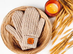 Beige crochet mittens with ribbed cable texture in a wooden bowl beside a candle and wheat stalks