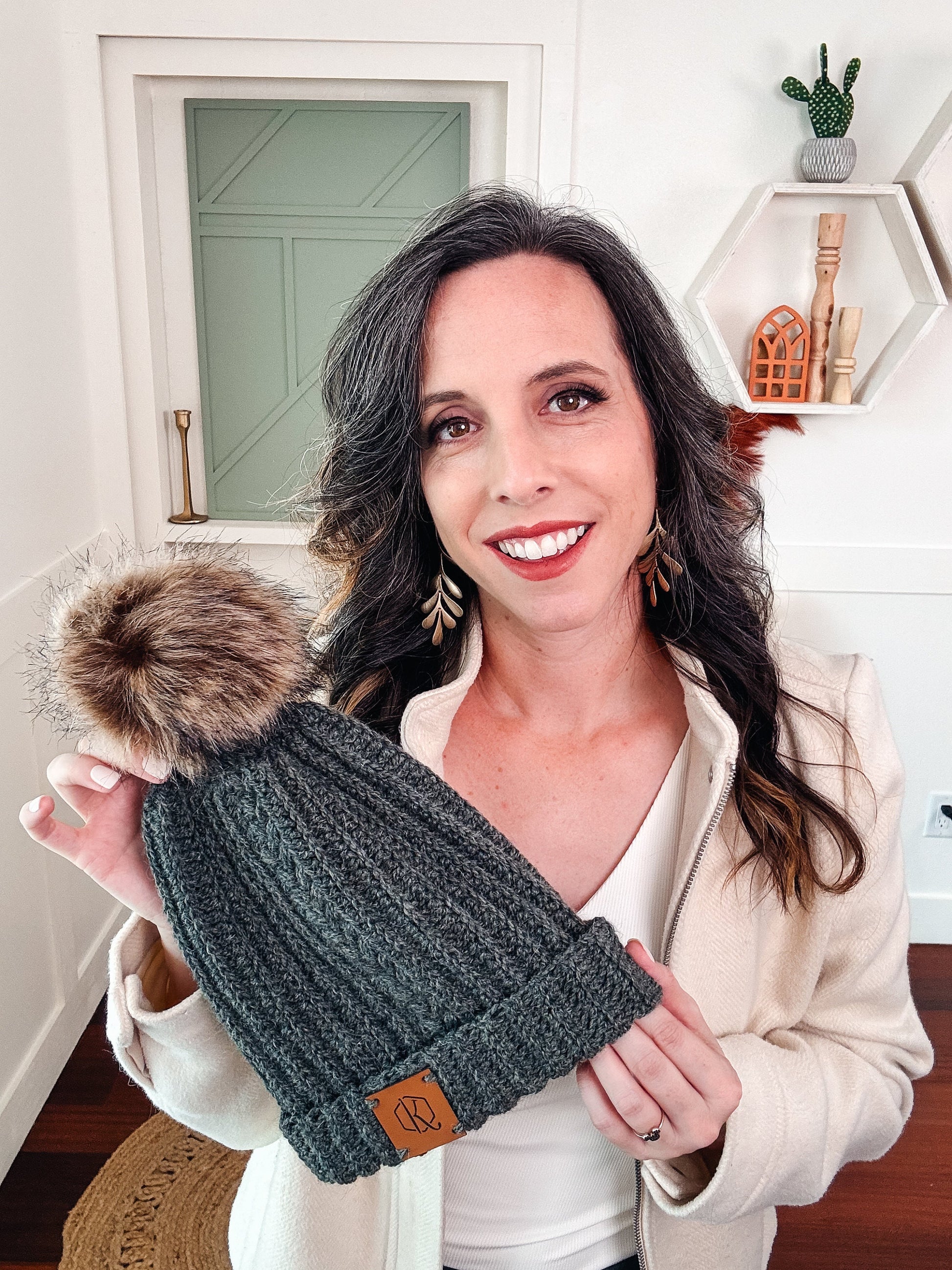 A woman smiles, holding a gray knit hat with a fur pom-pom, a charming handmade gift. She stands in a room with white walls and shelves displaying decor items.