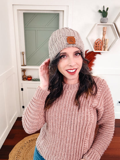 Taupe ribbed crochet beanie with a leather patch worn by a woman in a pink sweater
