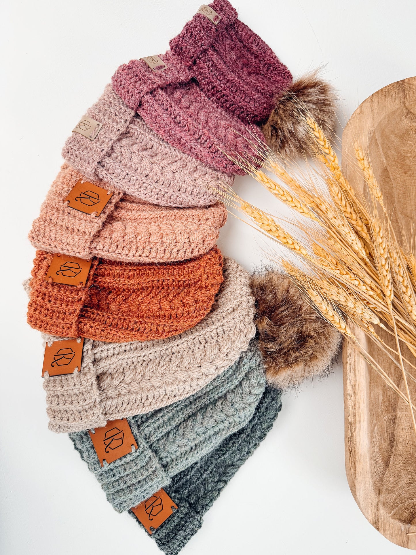 Stack of crocheted headbands in various colors with fur pom-poms on one end, perfect as a handmade gift, displayed alongside dried wheat stalks on a wooden tray.