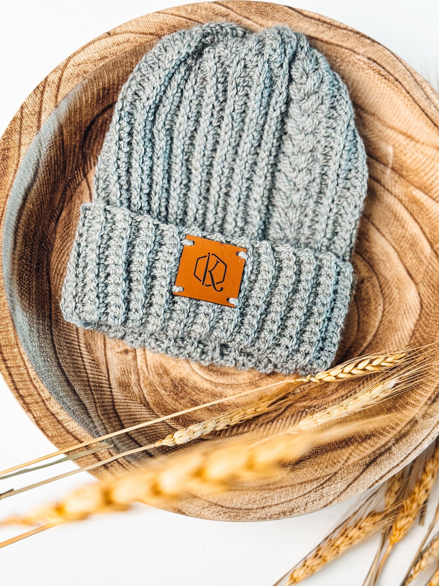 A gray knitted beanie with a leather patch lies on a wooden tray, surrounded by wheat stalks. This handmade gift adds a cozy touch to any winter ensemble.
