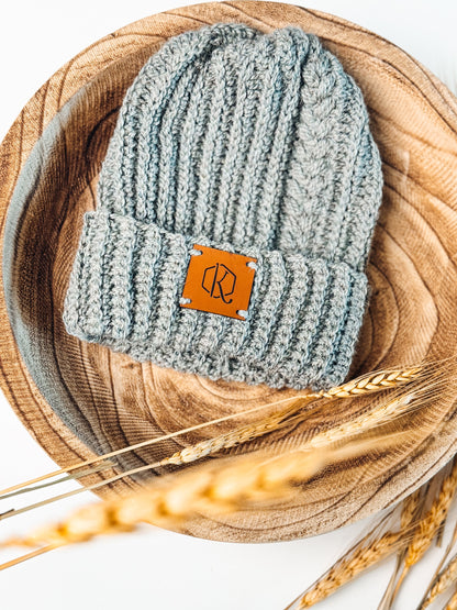 Light gray crochet beanie with braided texture and leather patch in a wooden bowl with wheat stalks