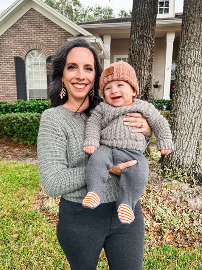 A woman holding a baby wearing a crochet sweater and hat in front of a unique house.