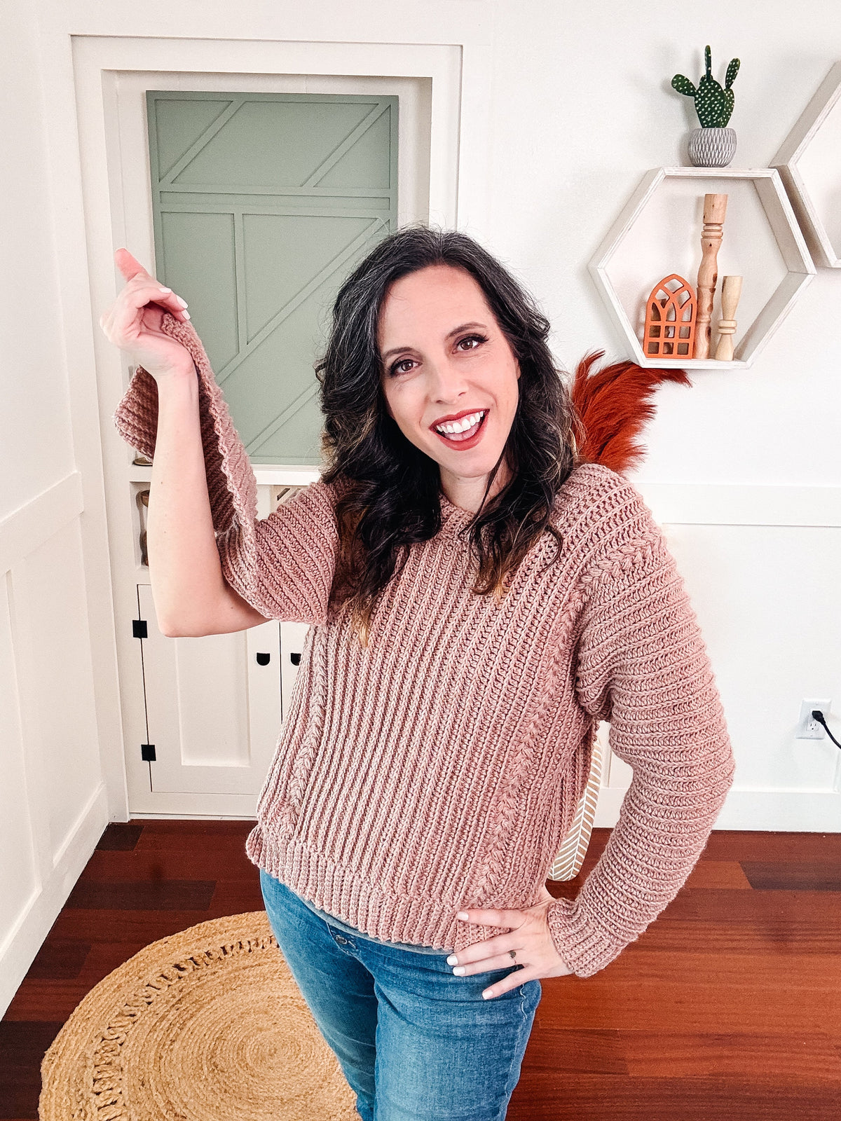 A woman wearing a crochet pink sweater and jeans standing in a room, accessorized with unique jewellery.
