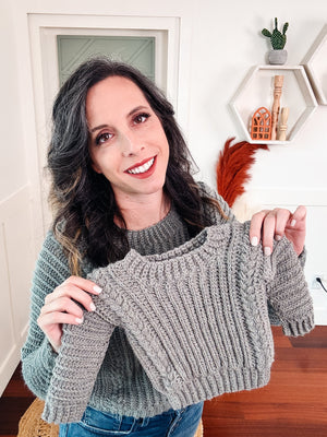 Smiling woman in a sage sweater holding up a small gray crochet child sweater with braided ribs