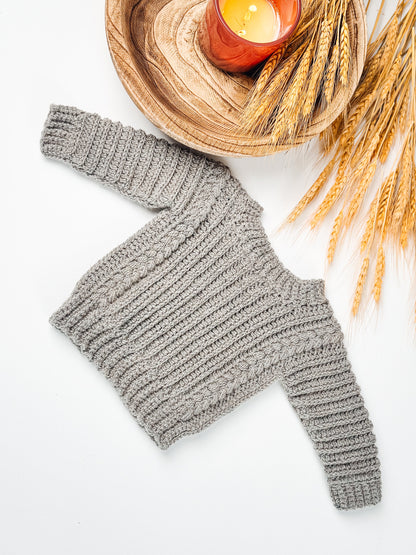 Autumn Wheat Child Sweater crochet pattern: gray crochet child sweater with ribbed and braided texture laid flat beside wheat stalks and a candle