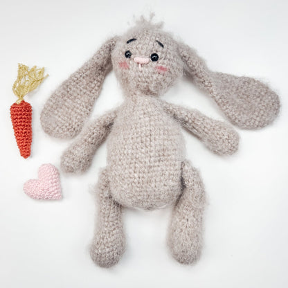 Tan fuzzy crochet bunny lying flat beside a small orange carrot and a pink heart on a white background
