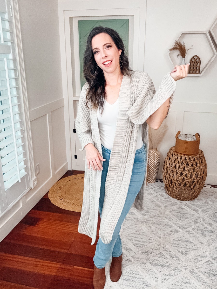 A woman stands in a well-lit room, smiling at the camera, wearing a white top, long beige crochet cardigan, blue jeans, and brown shoes adorned with unique jewellery.