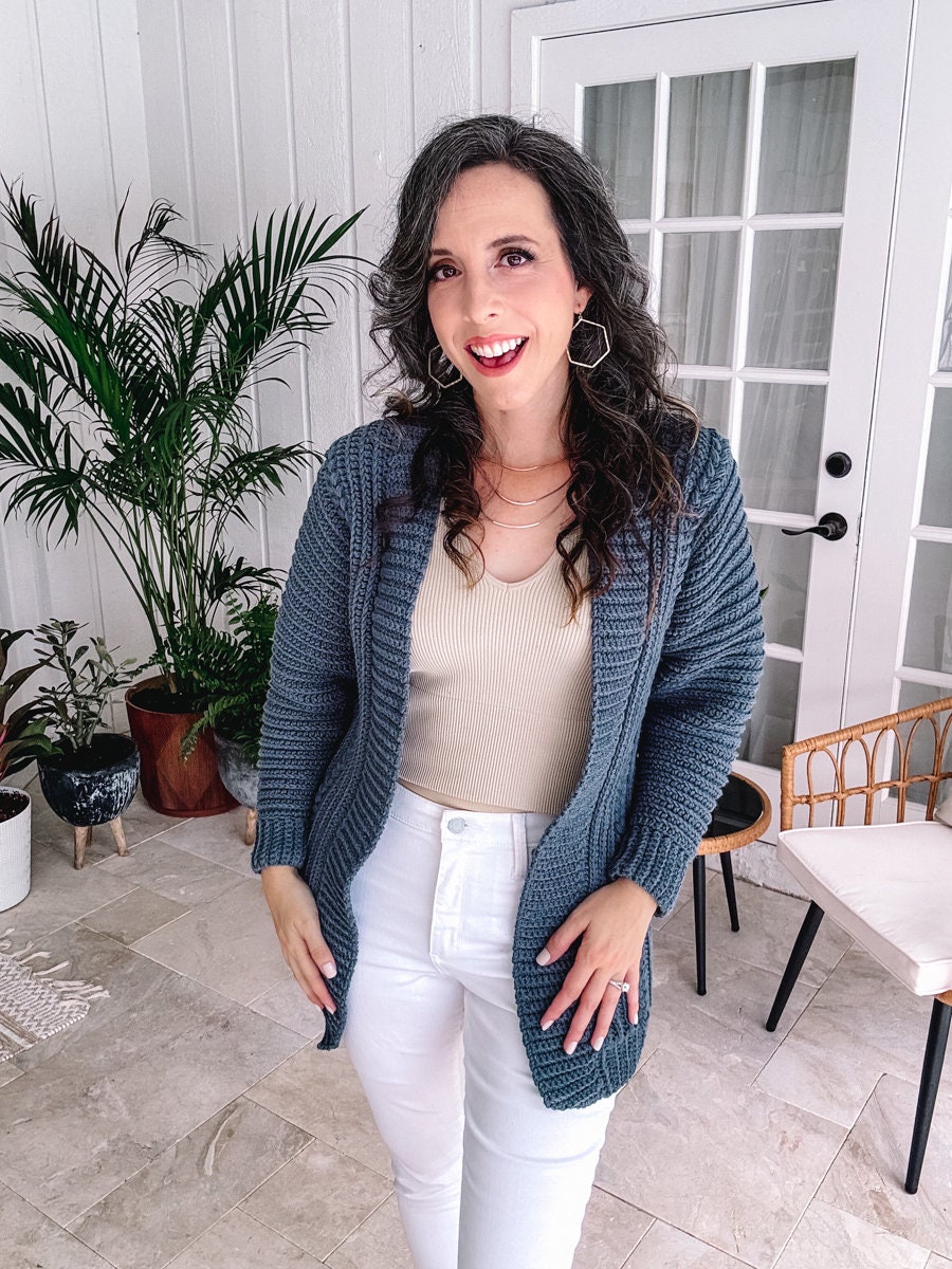 A person with long dark hair wearing a blue crocheted cardigan and white pants stands indoors near plants and furniture, adorned with unique jewellery.