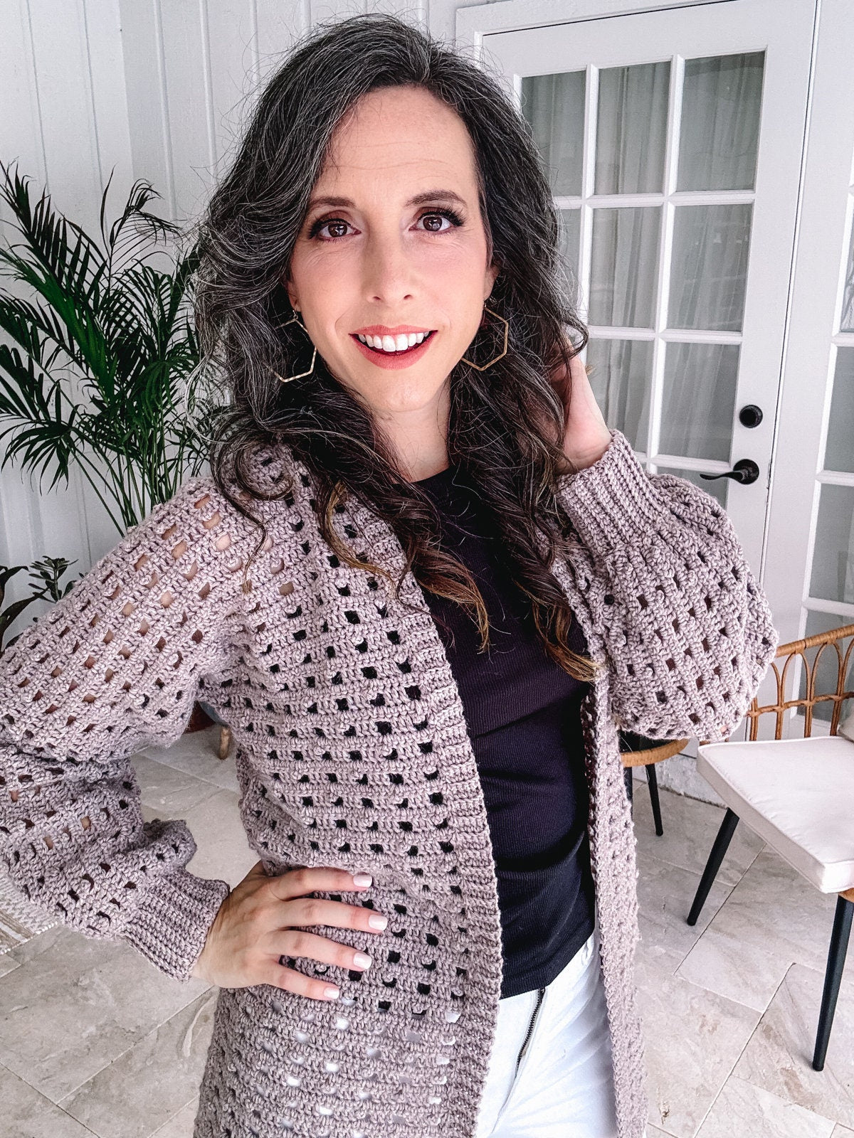 A smiling woman with curly hair wearing a beige crochet cardigan, black top, and white pants, standing indoors, accessorized with unique jewellery.