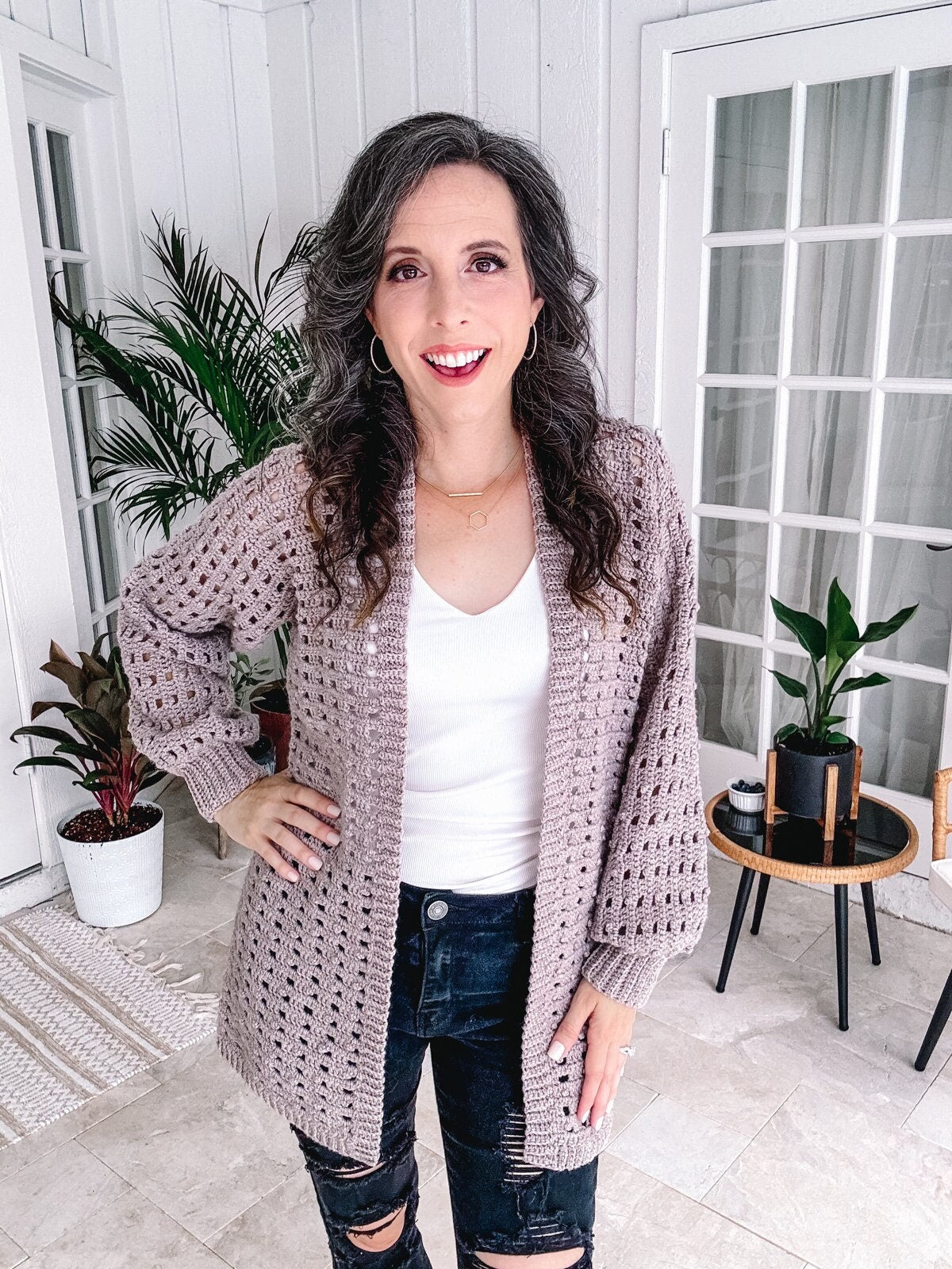 Woman standing in a room smiling, wearing a white top, a crochet cardigan, and ripped jeans with unique jewellery, with houseplants in the background.