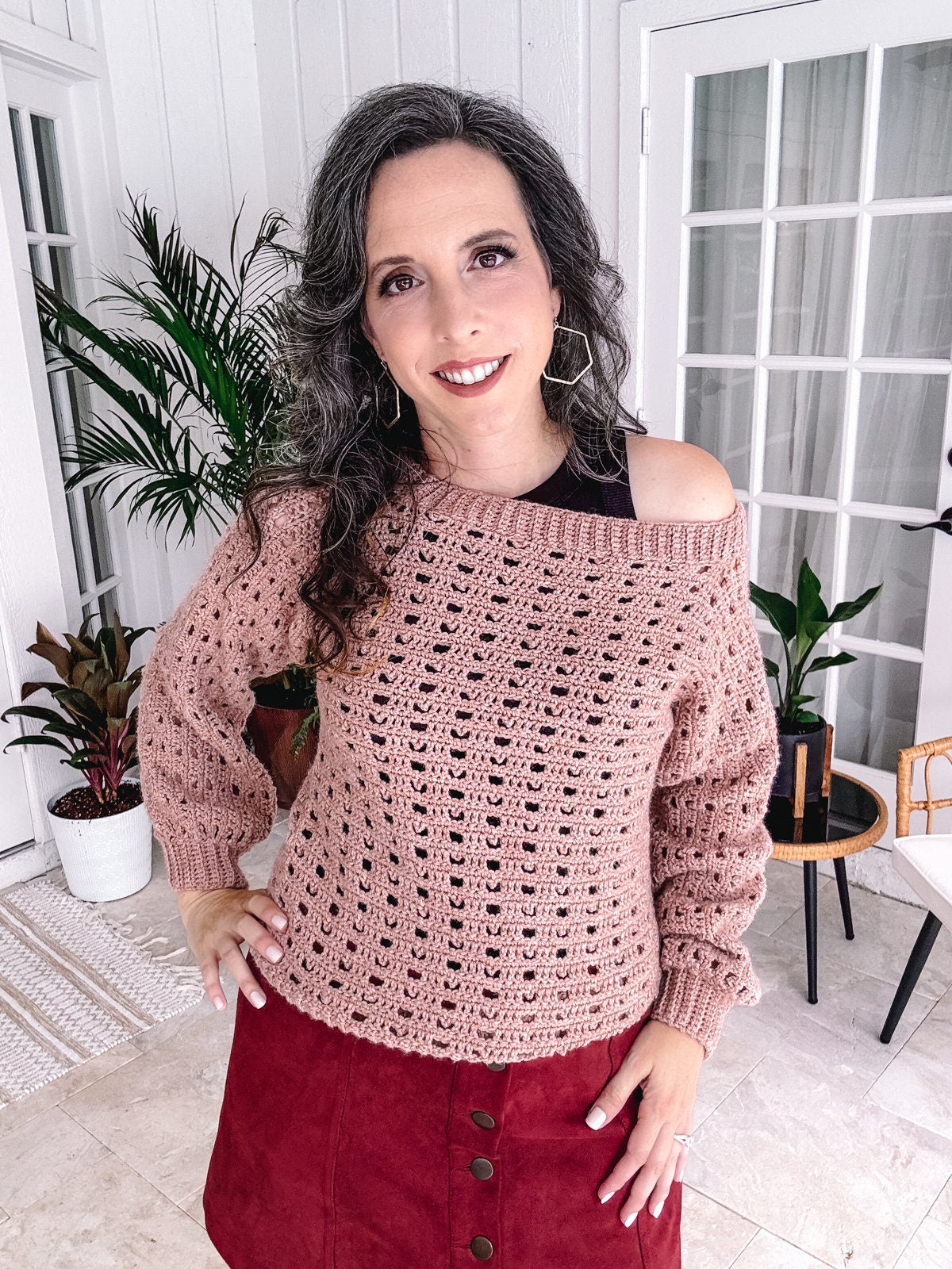 Woman posing in a room with an off-shoulder crochet top and a red skirt, adorned with unique jewellery.