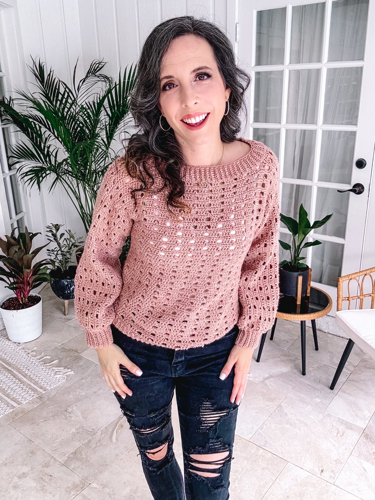Woman smiling in a room, wearing a pink crocheted sweater and black ripped jeans, accessorized with unique jewellery.