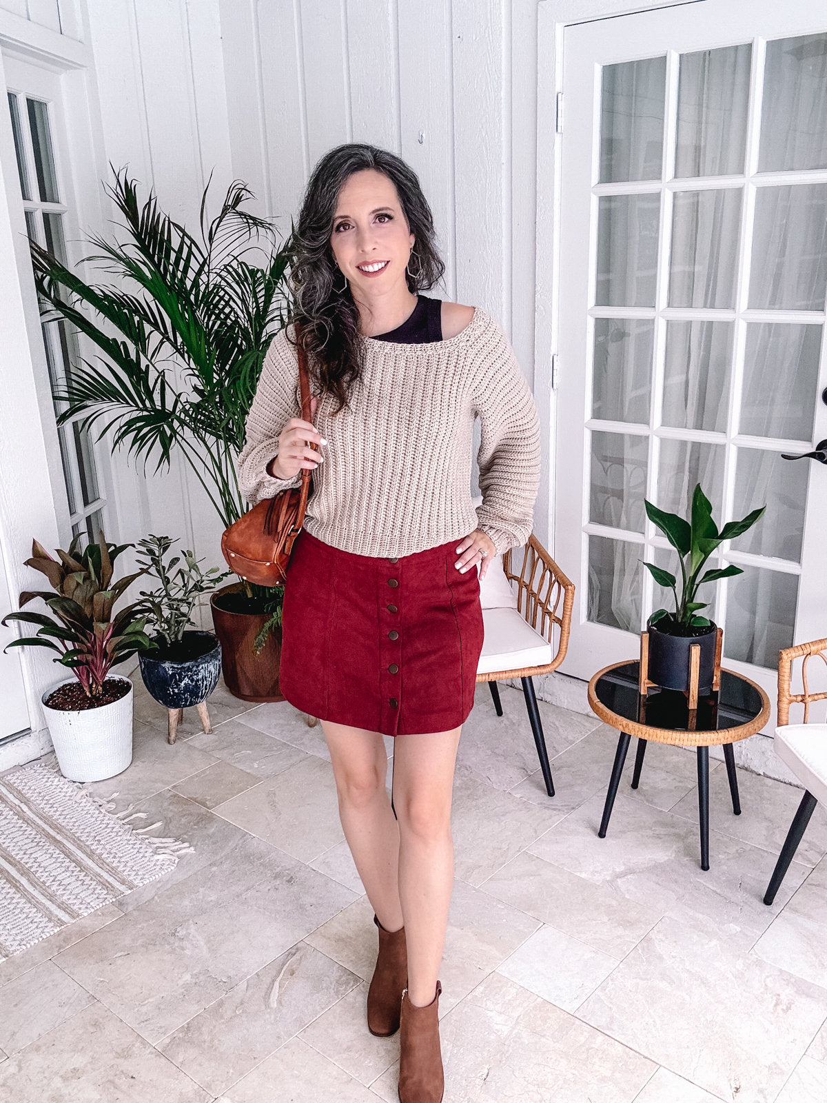 A woman standing indoors, smiling at the camera, dressed in a beige crochet sweater, red skirt, and brown boots, holding a handmade gift, with plants and furniture in the background.