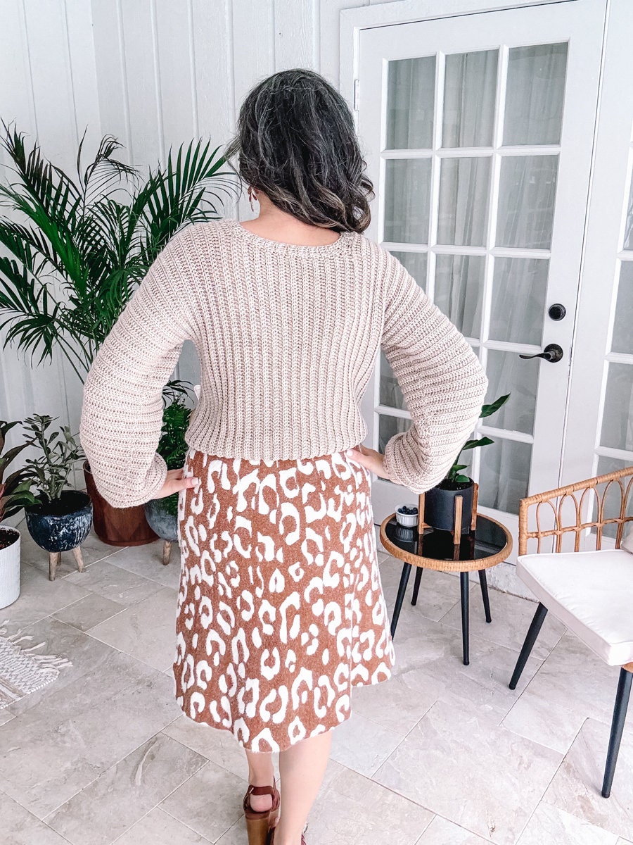 A person standing in a room with hands on hips, facing away from the camera, wearing a beige crochet sweater and a leopard print skirt paired with unique jewellery.
