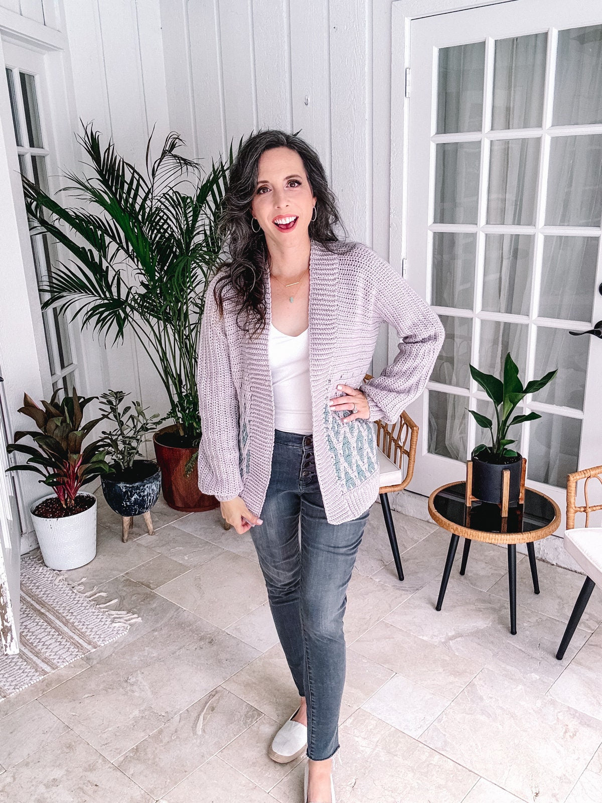 A woman standing indoors with one hand on her hip, wearing a white top, gray cardigan, and denim jeans, surrounded by houseplants and unique jewellery.