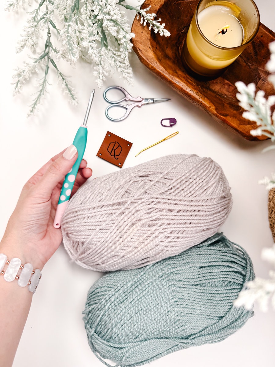 A person holds a crochet hook beside two balls of yarn, scissors, a candle, and decorative plants on a white surface to create unique jewellery.