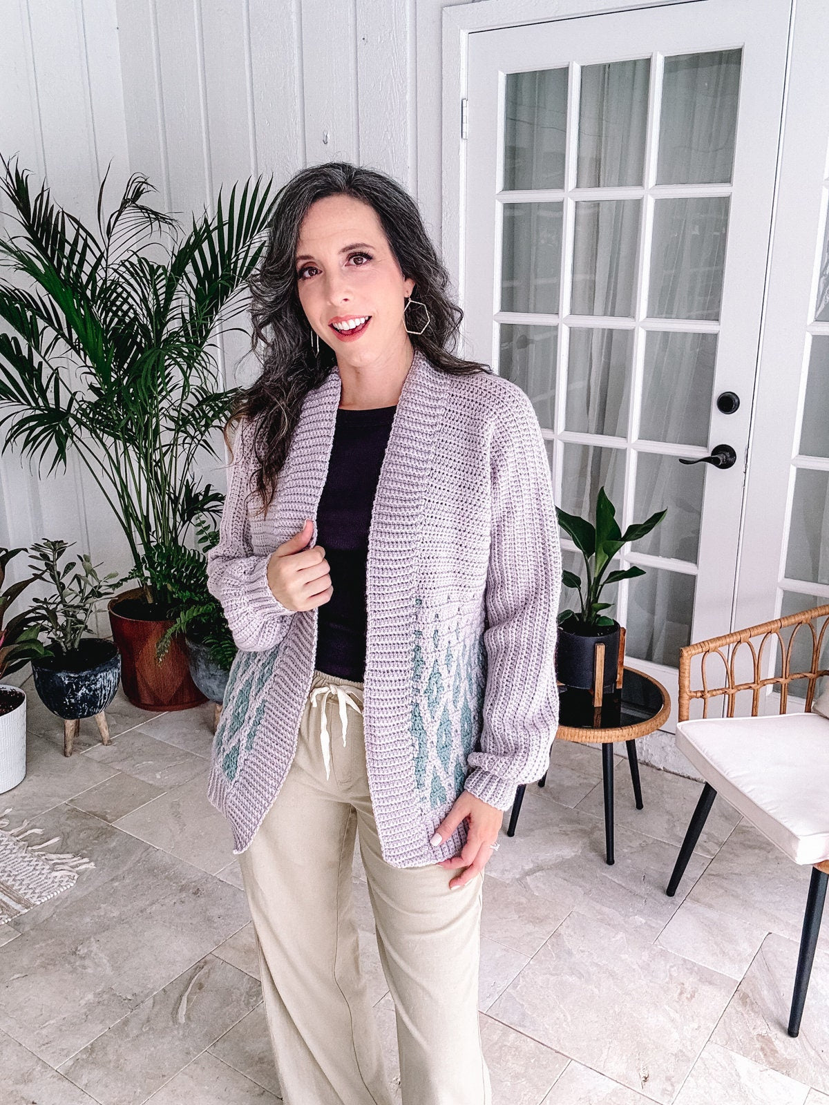 A woman in a crochet cardigan and casual trousers standing indoors with potted plants in the background, wearing unique jewellery.