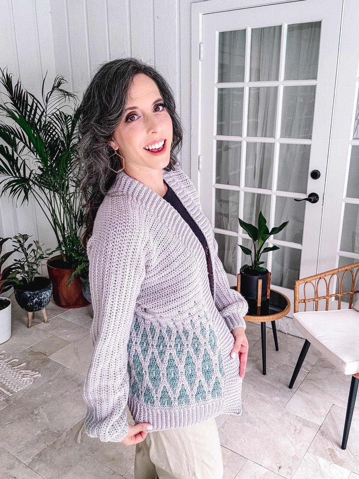 Woman standing indoors wearing a crochet purple sweater and beige pants, smiling at the camera with her hand on her hip.