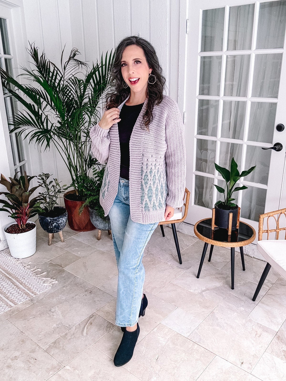 Woman posing indoors holding the edge of a cardigan, dressed in a casual chic outfit including a patterned cardigan, jeans, and ankle boots, surrounded by houseplants.