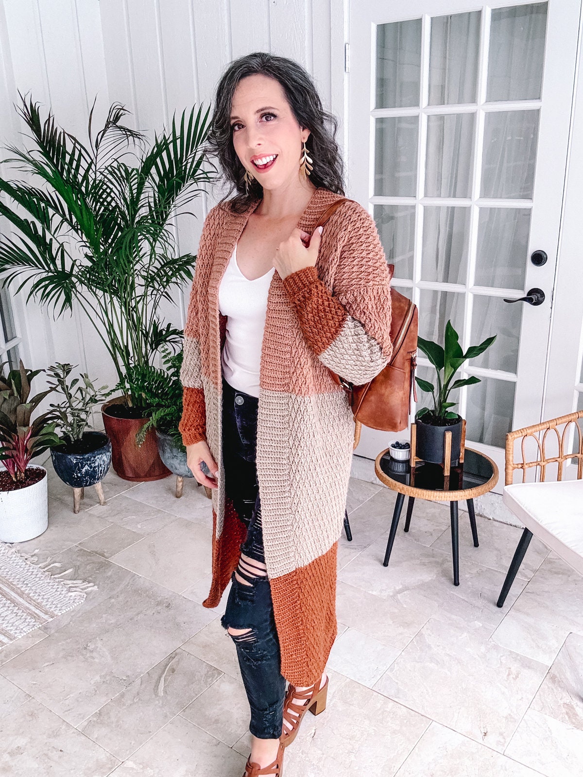 A woman in a cozy, oversized crochet cardigan stands smiling in a stylishly decorated room, wearing unique jewellery.