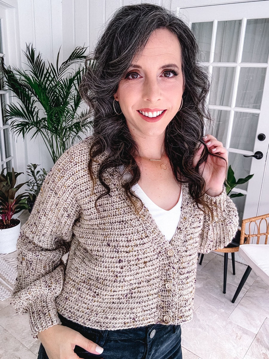 A person with curly hair smiling at the camera, wearing a beige cardigan and a white top, indoors with plants in the background, and showcasing unique jewelry.