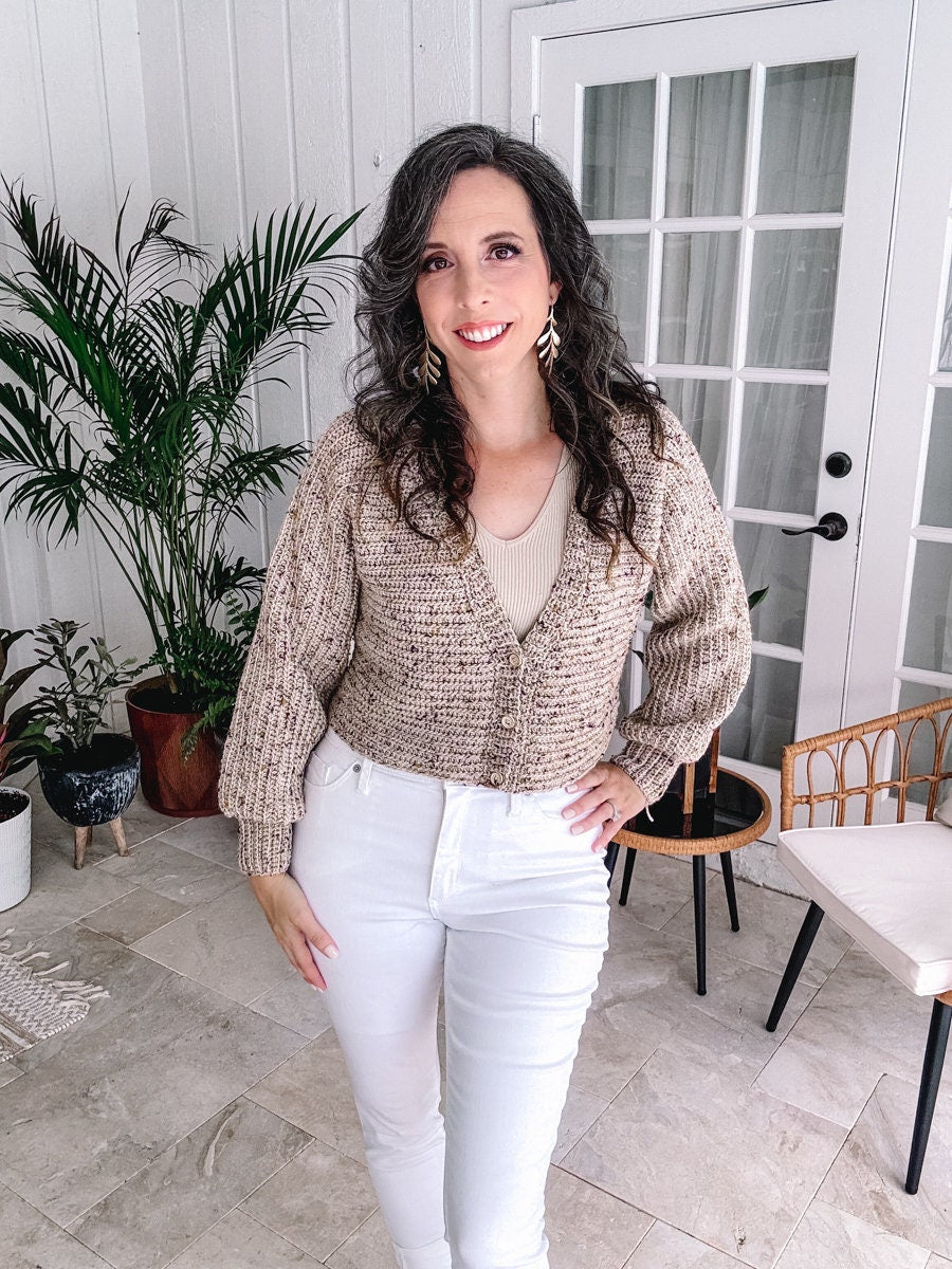A woman with curly hair smiling indoors while wearing a beige cardigan, white pants, and a light top adorned with unique jewellery.