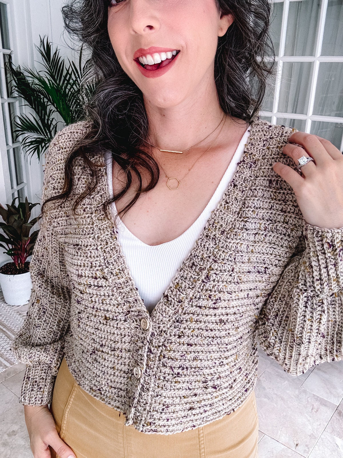 A person wearing a textured cardigan, white top, and unique jewellery, standing indoors.