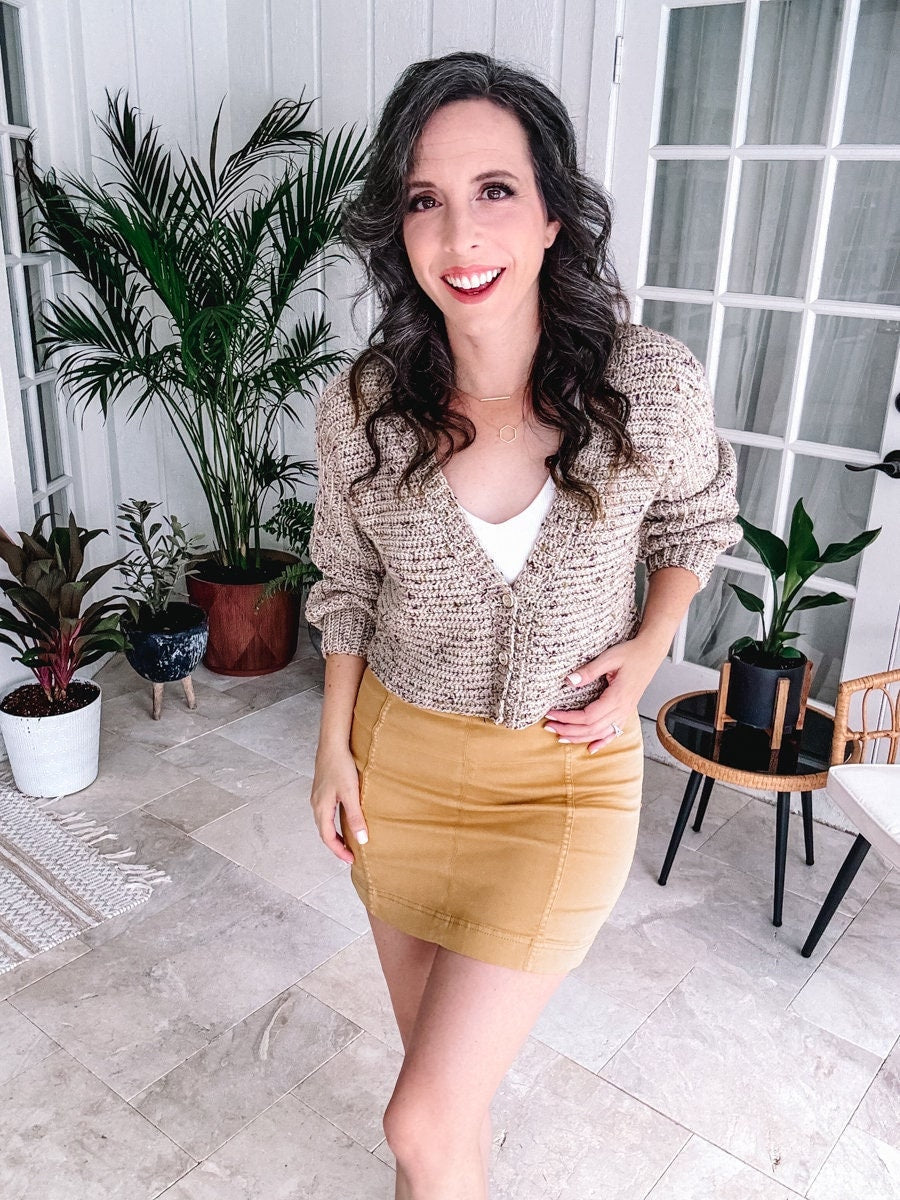 A woman smiling in a beige crochet cardigan and mustard skirt, accented with unique jewellery, standing in a room with potted plants.