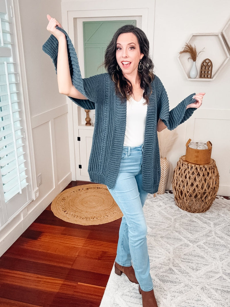 Slate blue crochet cardigan held out at the sleeves by a smiling woman in a white top and light blue jeans