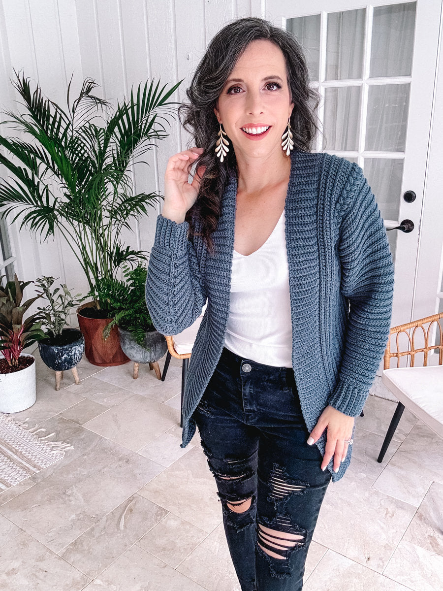 A person indoors, styled in a cochet blue cardigan, white top, and black ripped jeans, stands near plants and a chair. Their look is elevated with unique jewellery that adds a touch of elegance to their ensemble.