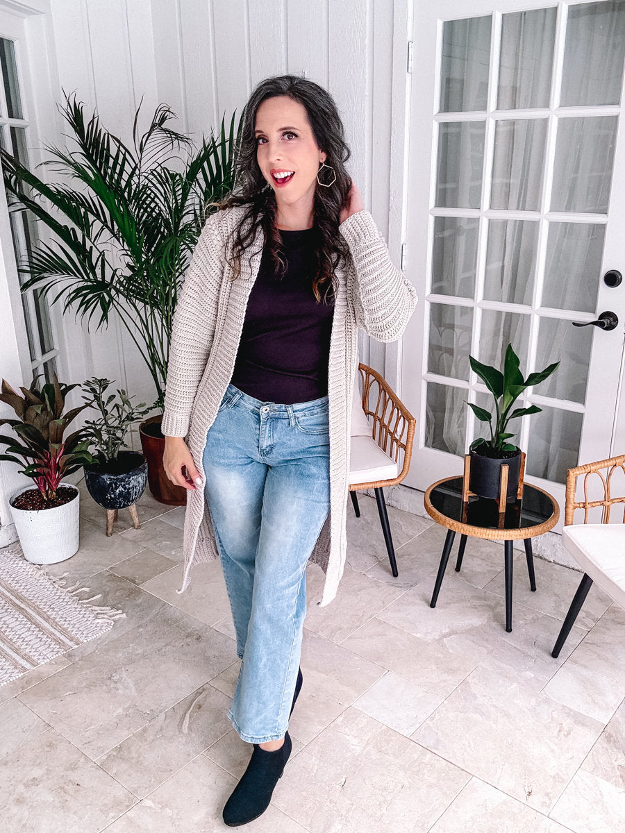 A person with long hair is standing indoors near plants, donning unique jewellery that complements their long beige cardigan. They sport a dark top, light blue jeans, and black shoes. The scene features a tiled floor and glass doors in the background.