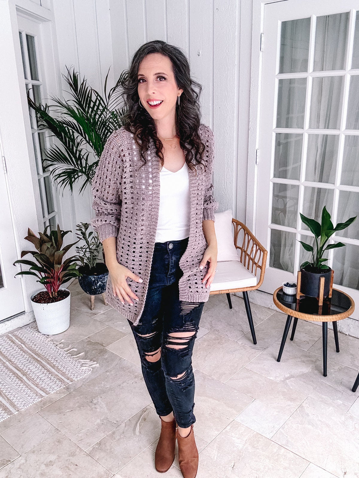 Woman in a casual outfit with ripped jeans and a handmade crochet cardigan standing in a stylish home interior.