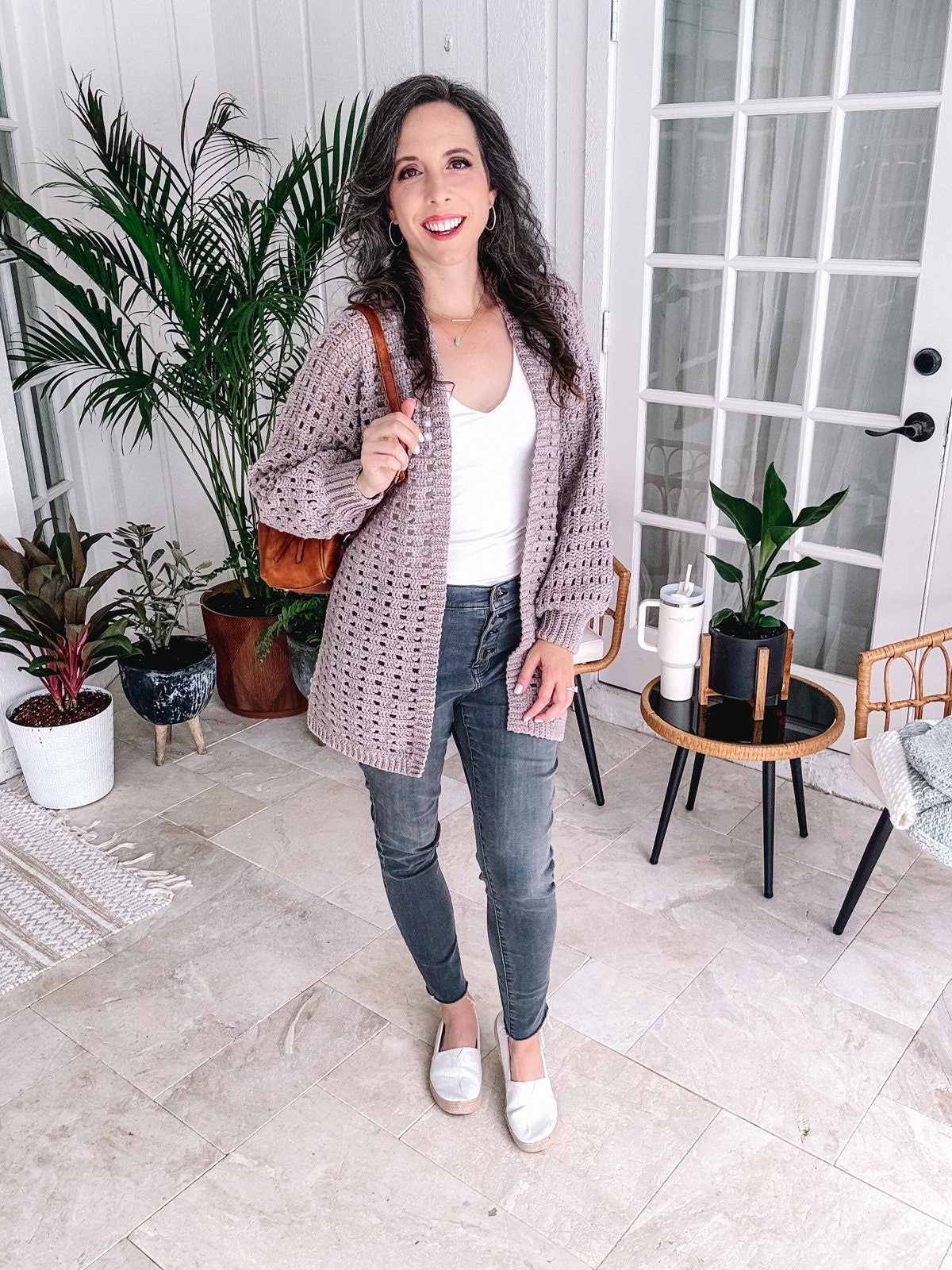 A woman in a white top, grey jeans, and a light crochet cardigan stands smiling in a room with indoor plants and modern decor, showcasing her unique jewellery.