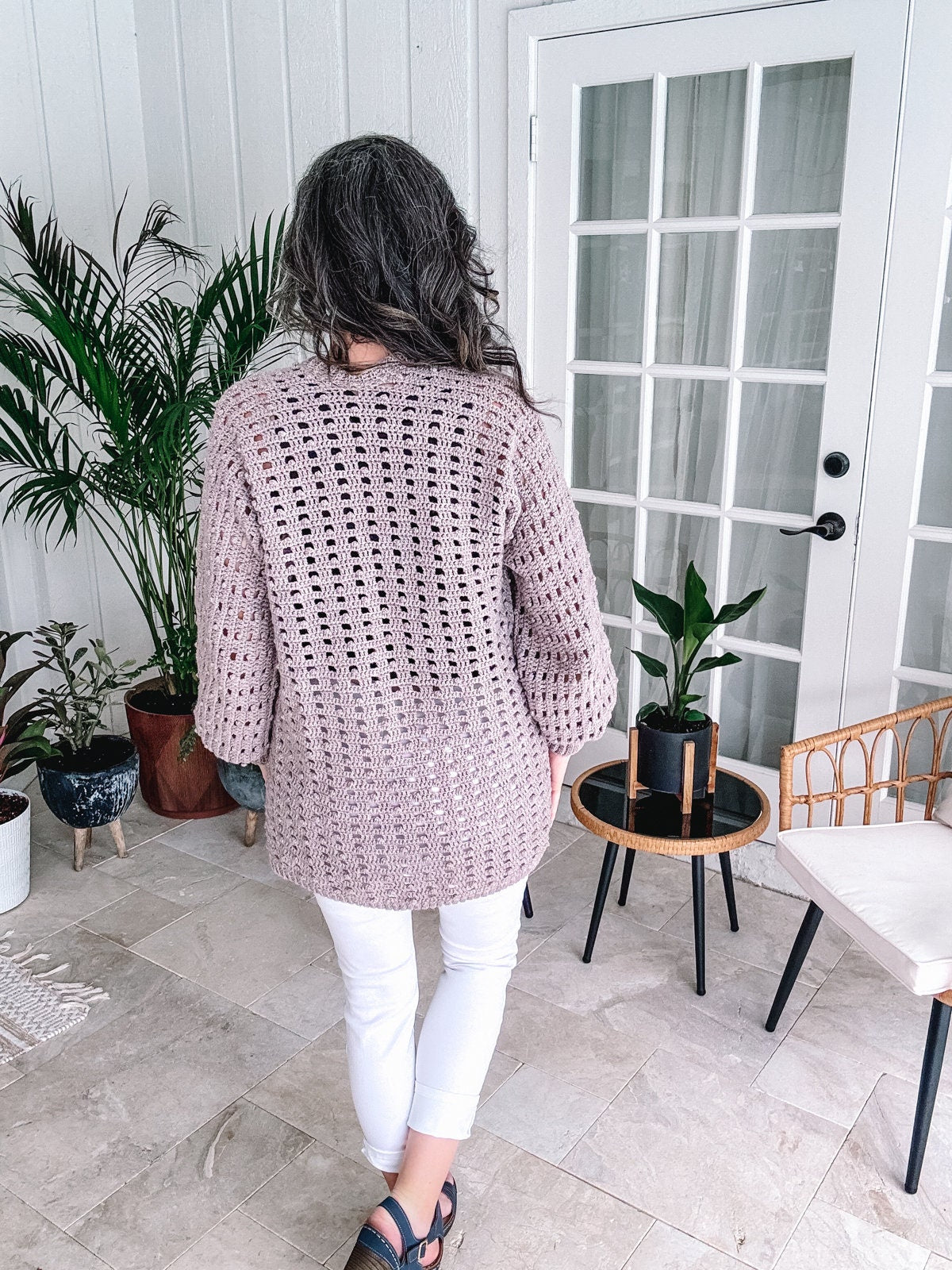 Woman standing on a patio wearing a loose-crochet sweater and white pants, facing away from the camera toward a door flanked by plants, showcasing her unique jewellery.
