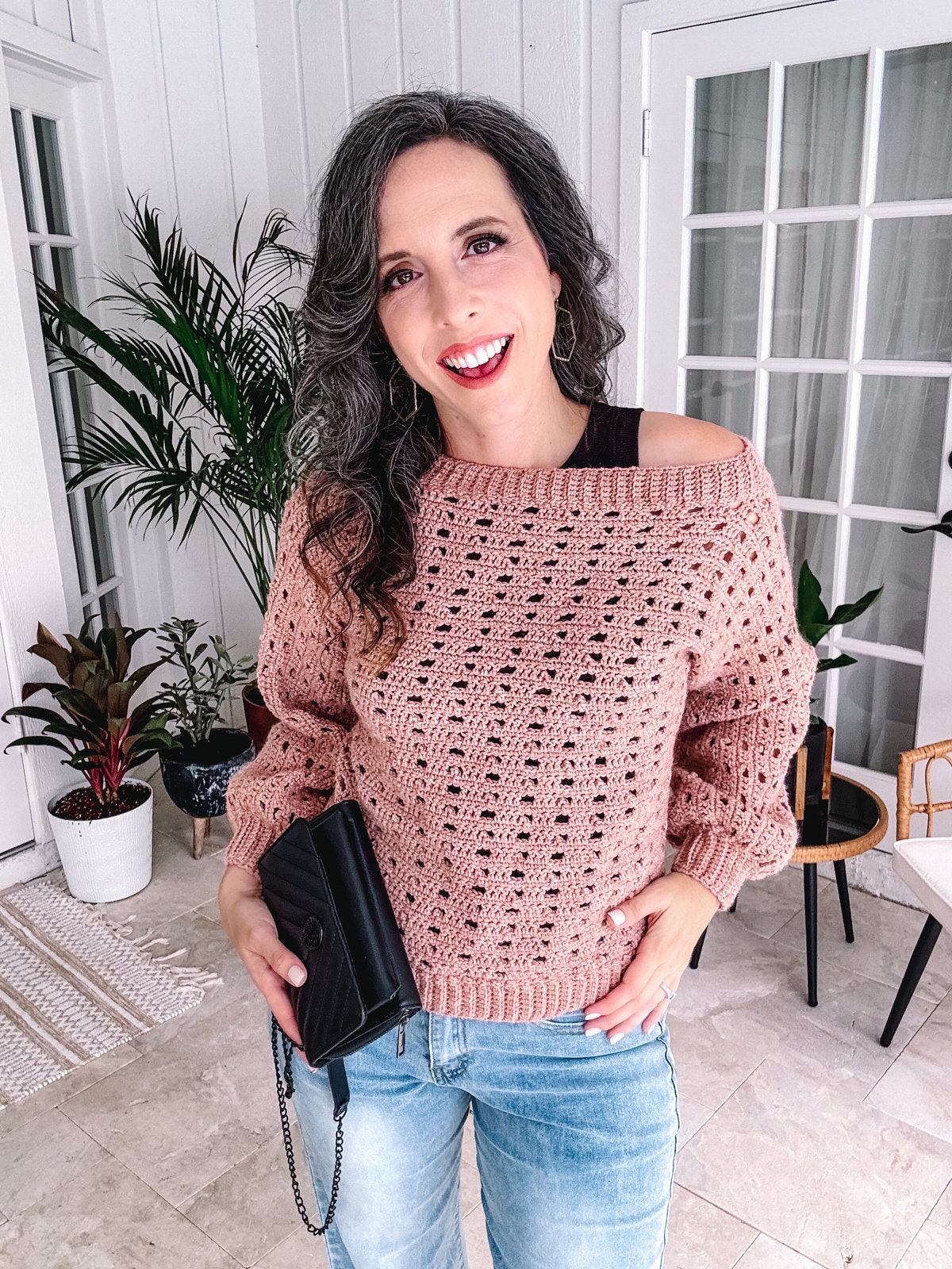 Woman smiling indoors wearing an off-shoulder crochet sweater and jeans, holding a black purse and unique jewellery.