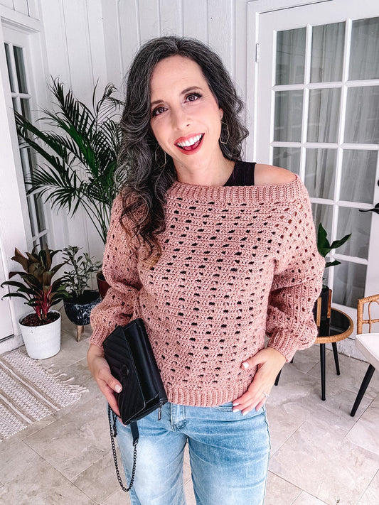 Woman smiling indoors wearing an off-shoulder crochet sweater and jeans, holding a black purse and unique jewellery.