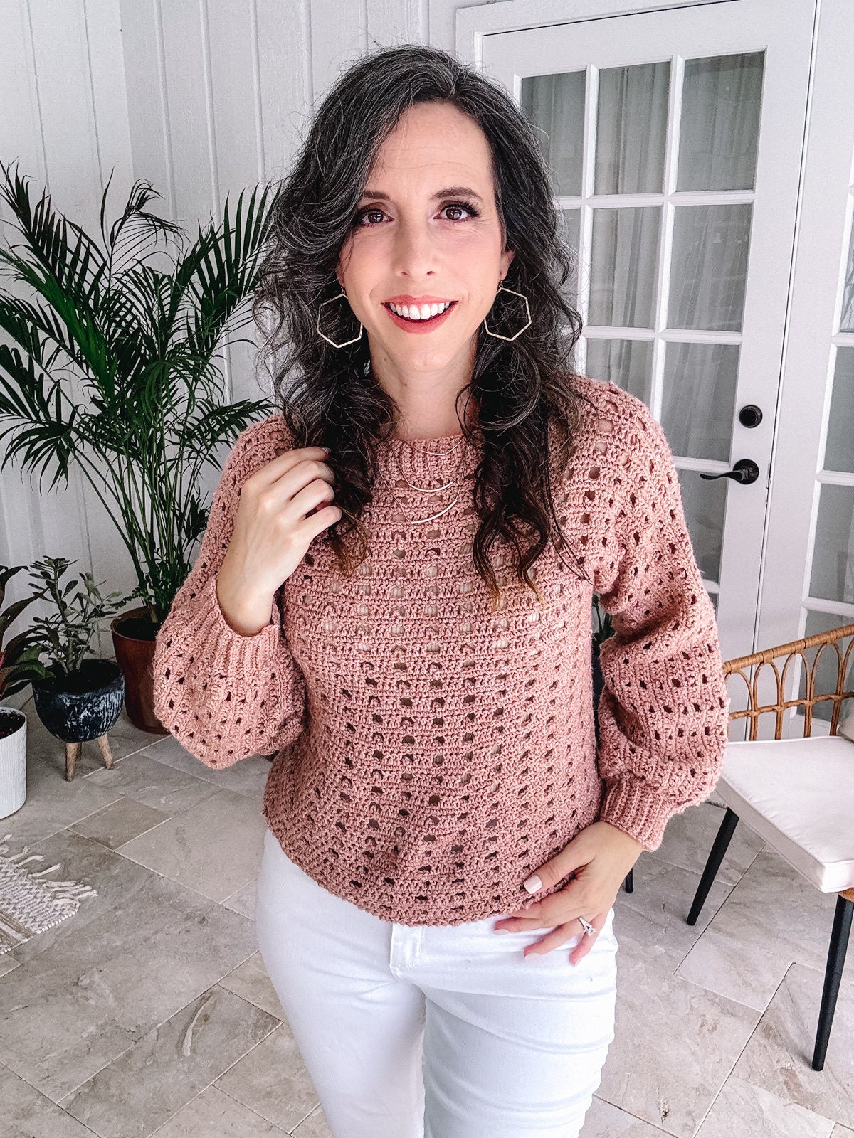 Woman in a vintage pink crochet sweater and white pants standing in a room with a plant and French doors in the background.