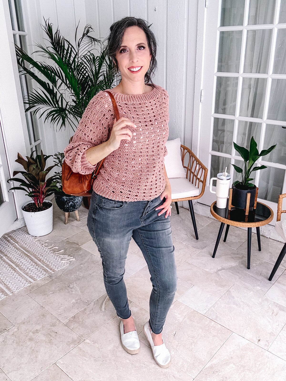 A woman in a pink crochet top and gray jeans standing indoors, accessorized with a leather shoulder bag, white loafers, and unique jewelry.