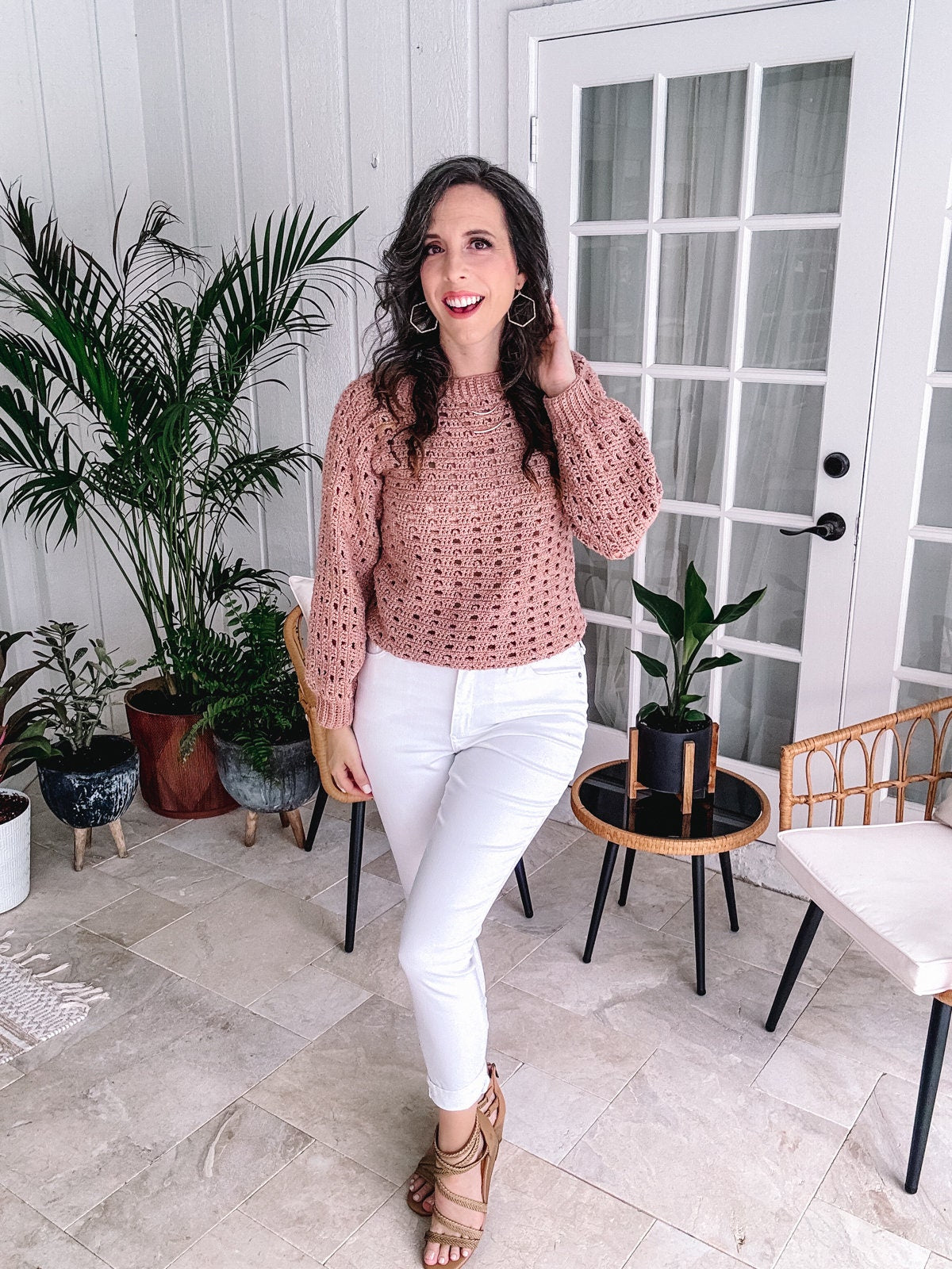 A woman standing on a patio wearing a crochet top, white jeans, and strappy sandals with unique jewellery and indoor plants in the background.
