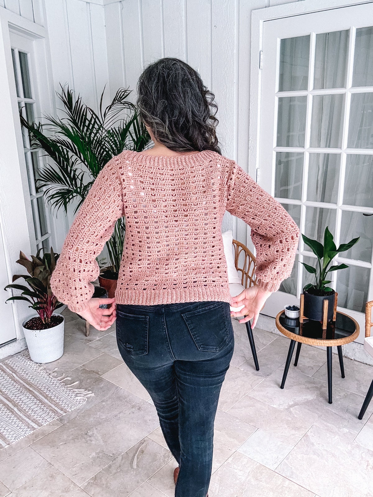A woman from behind wearing a unique jewellery piece, a pink crochet sweater, and blue jeans in a room with houseplants.