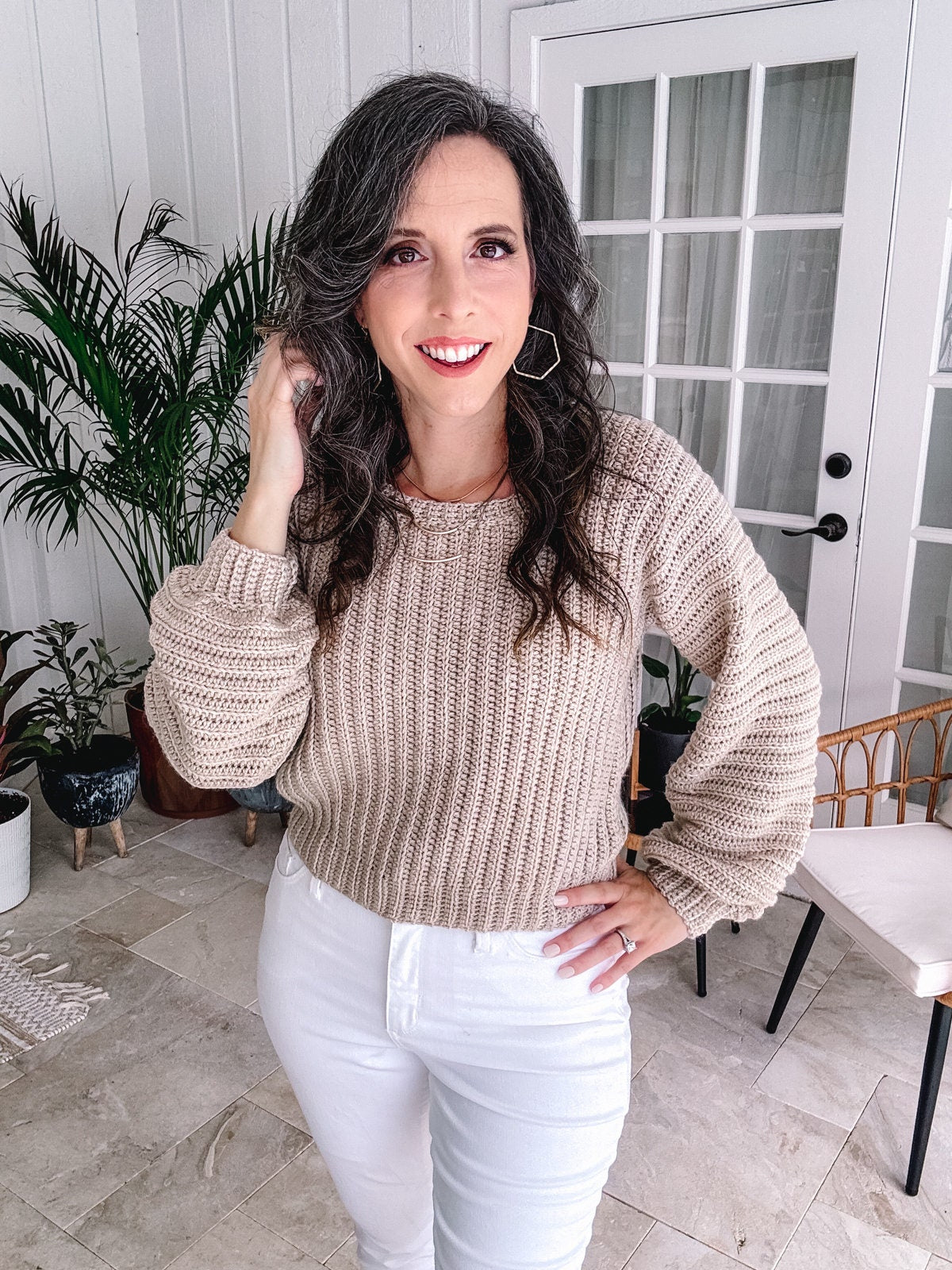 Woman posing with hand in hair, wearing a beige crochet sweater and unique jewellery, in a room with plants and French doors.