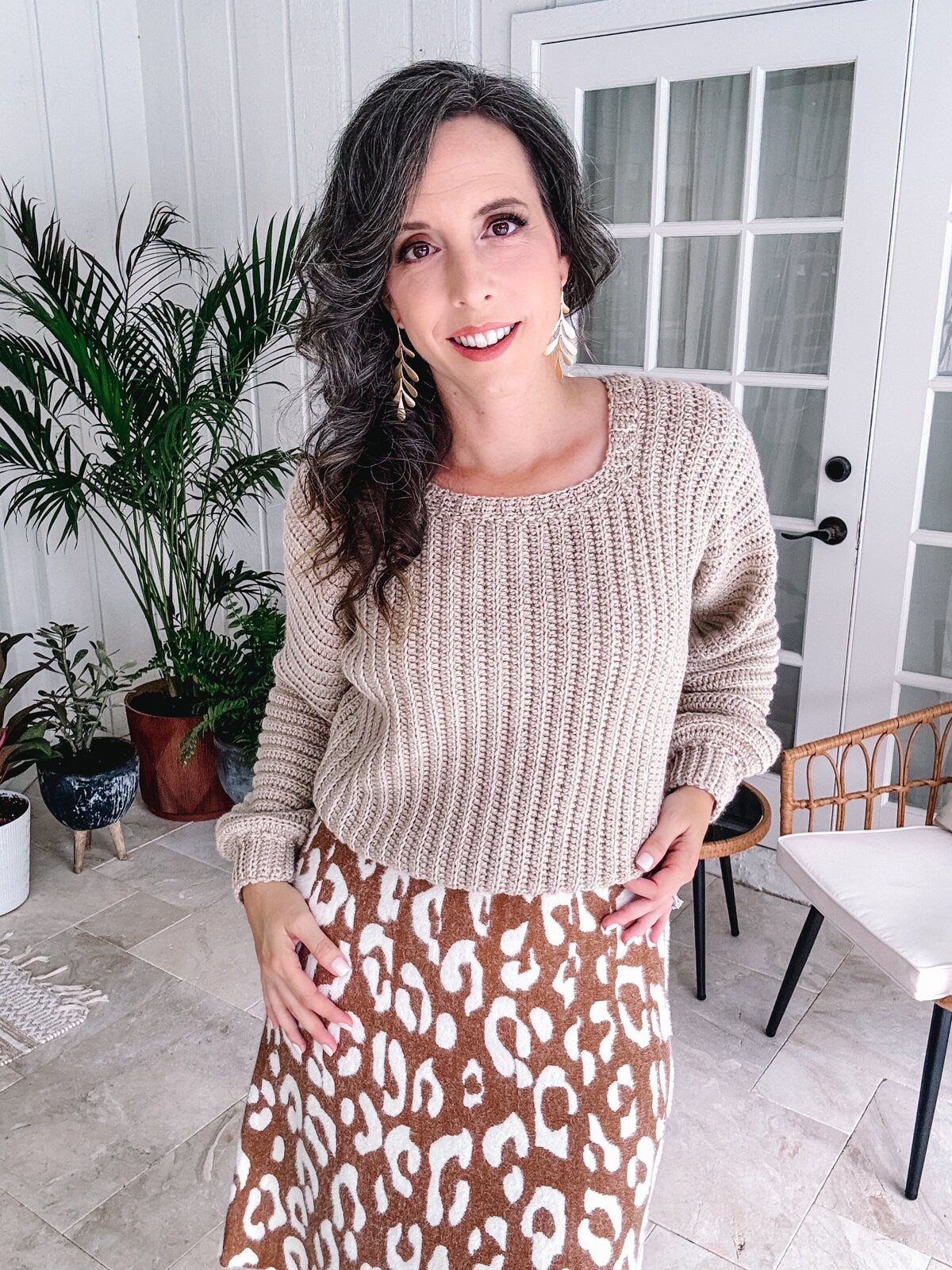 Woman in a beige crochet sweater and animal print skirt, wearing unique jewellery, smiling in a well-lit room with plants and furniture.