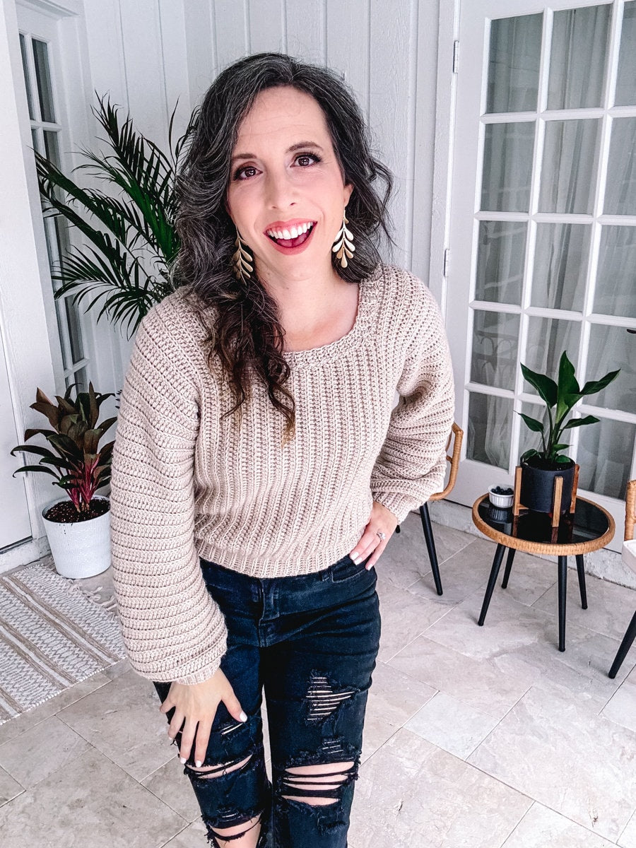 A woman with curly hair, wearing a crochet beige sweater and ripped jeans, smiles brightly indoors near potted plants, showcasing her unique jewelry.