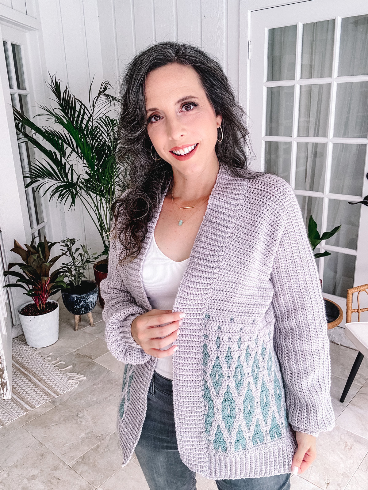 Woman in a white top and textured crochet cardigan standing in a room with plants, adorned with unique jewellery.