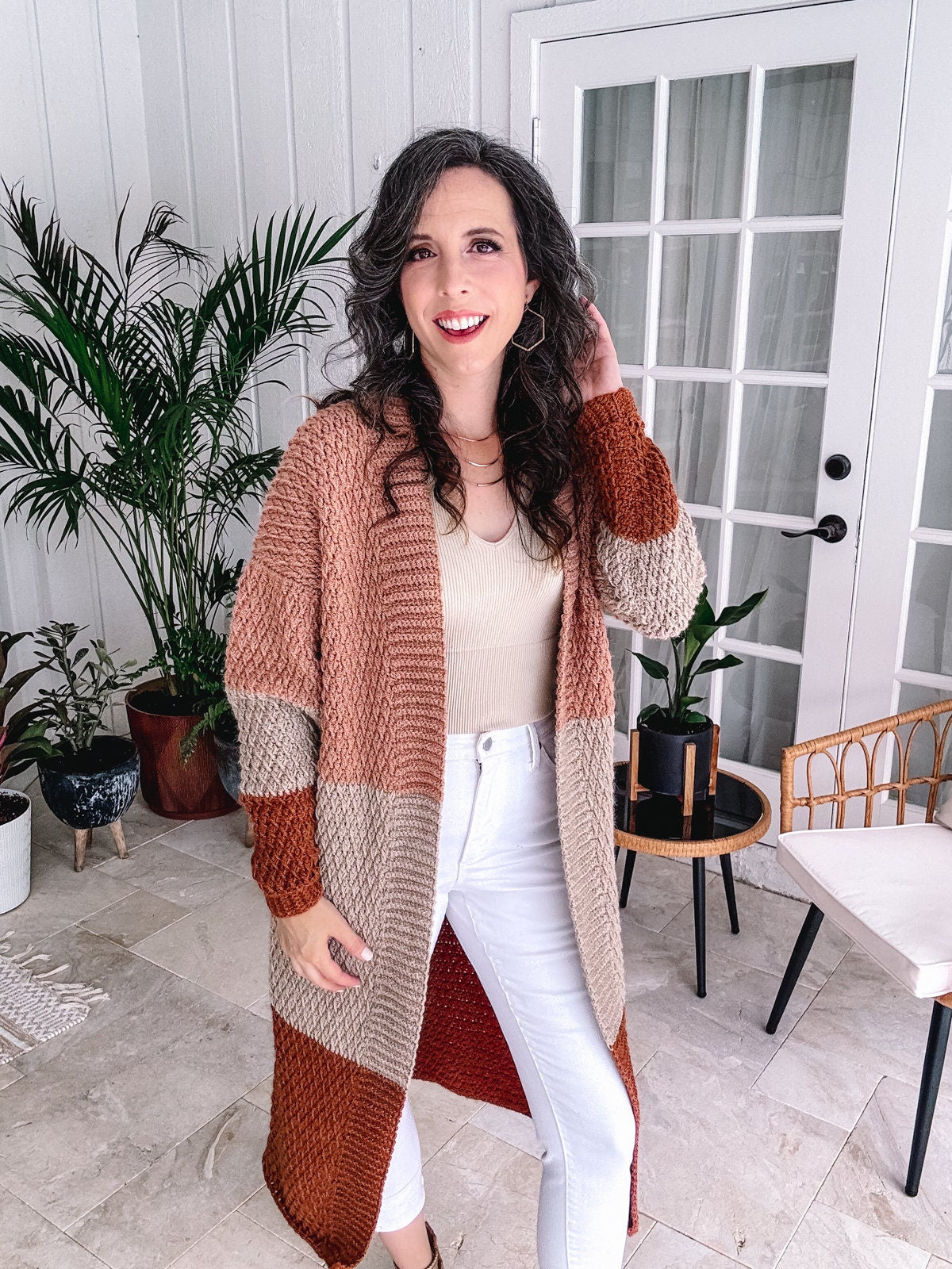 A smiling woman standing in a stylish interior, wearing a long, color-block cardigan, a beige top equipped with unique jewellery, and white pants.