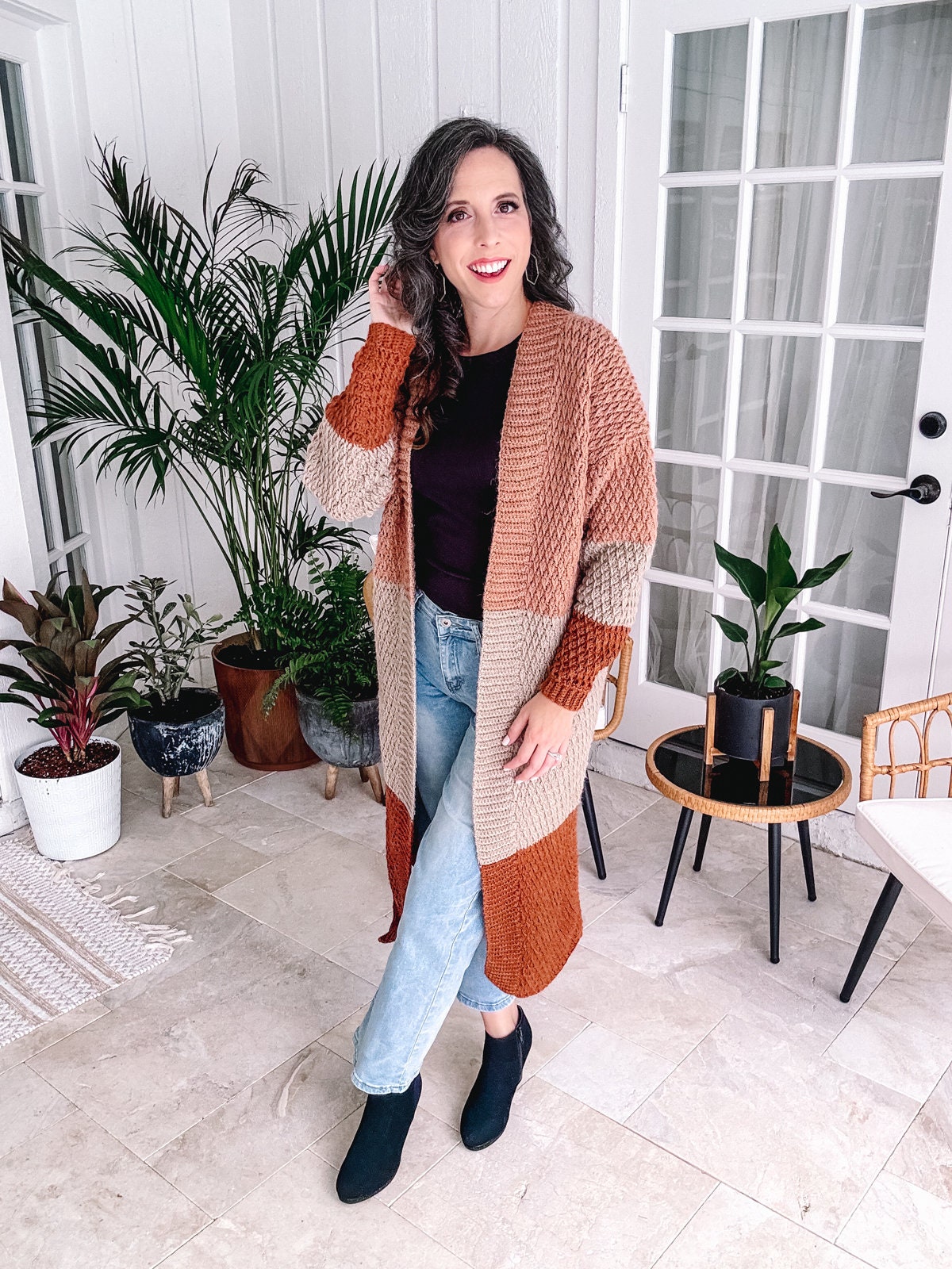 A woman in a stylish cardigan and jeans standing in a well-decorated room with houseplants, wearing unique jewellery.