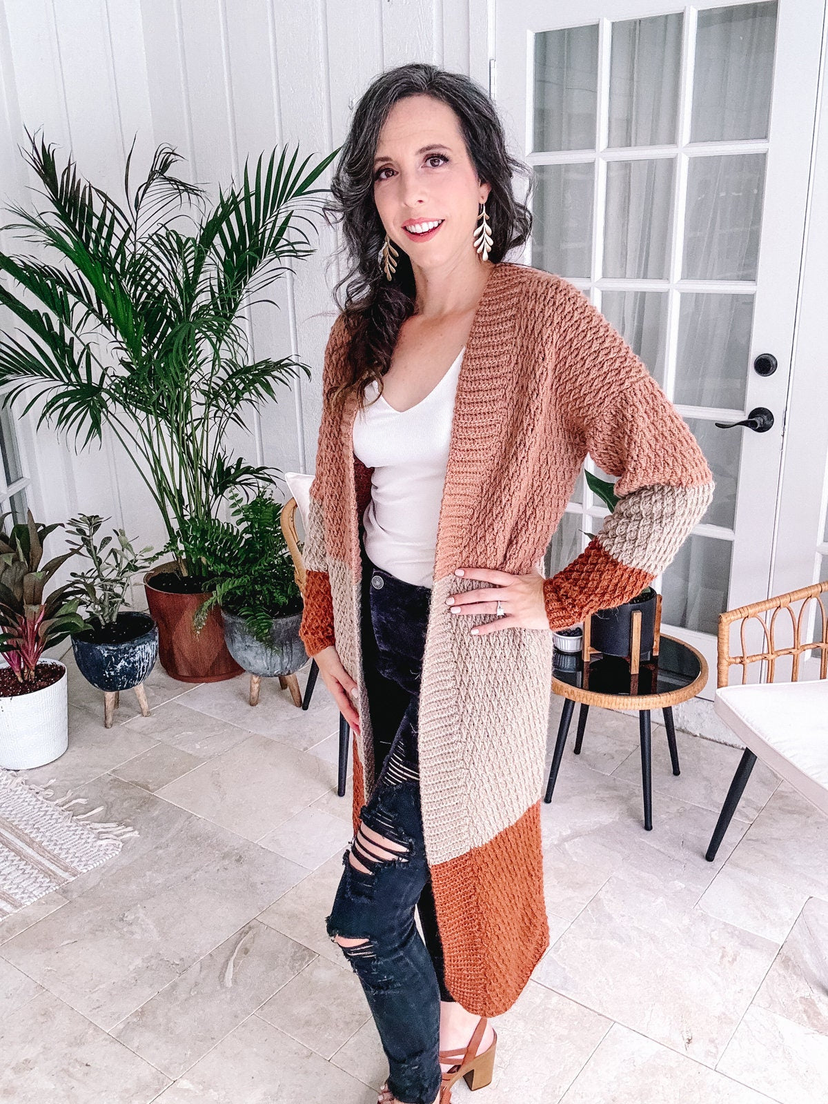 Woman posing in a stylish interior, wearing a unique jewellery piece with her color-blocked cardigan, ripped jeans, and tan heels.