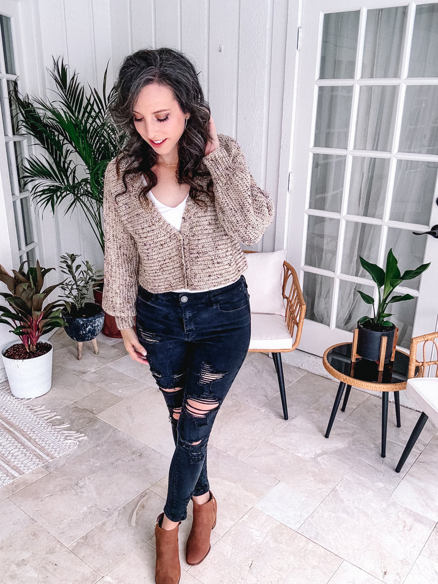 A person standing in a stylish indoor setting, dressed in a beige jacket, white top, ripped dark jeans, and brown boots, adjusting their hair while wearing unique jewellery.