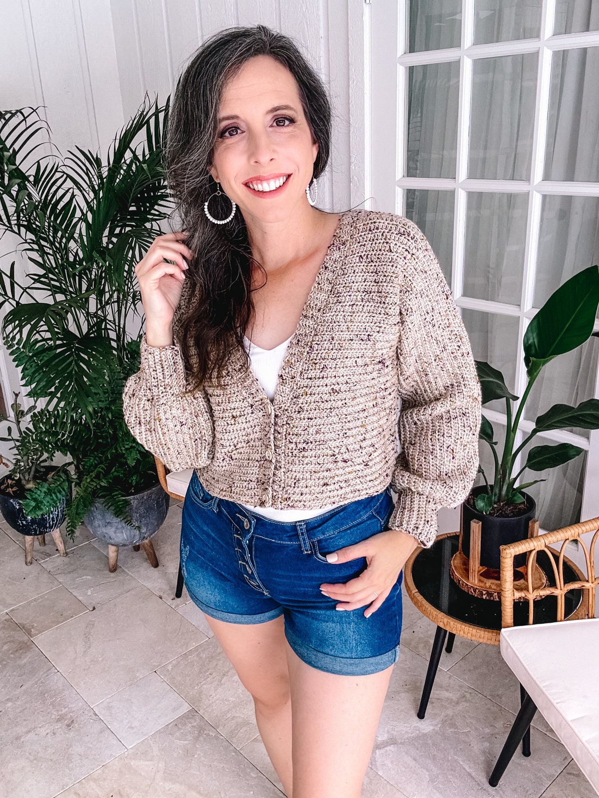 A woman smiling at the camera, wearing a knit cardigan and denim shorts, standing in a room with plants and a wicker chair, complemented by unique jewellery.